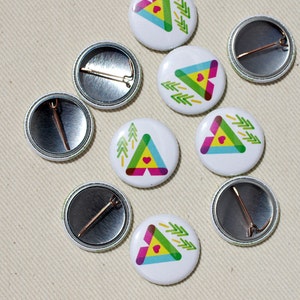 May include: Nine round white button pins with a colorful geometric design of a triangle with a heart inside. The design also includes green pine trees. Some of the pins have a silver back with a safety pin.
