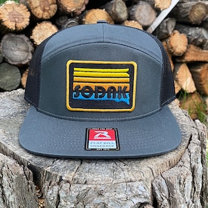 May include: A gray and black trucker hat with a black and yellow patch that says "SOPAK" in a retro font. The patch has a yellow, orange, and blue design that resembles a sunset.