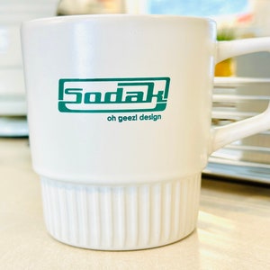 May include: White ceramic mug with a green logo that says "Sodak" and "oh geezl design". The mug has a ribbed design on the bottom.