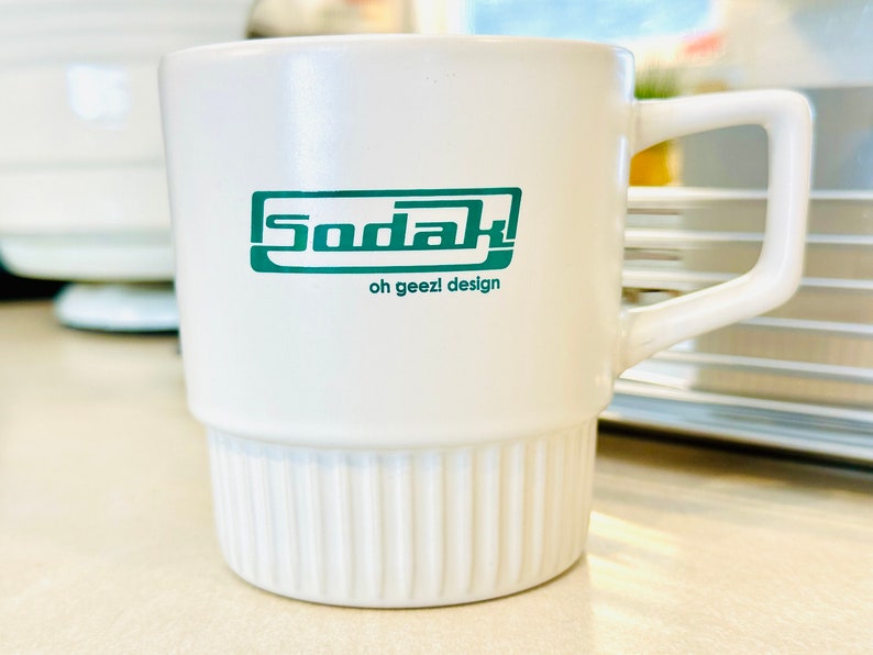 May include: White ceramic mug with a green logo that says "Sodak" and "oh geezl design". The mug has a ribbed design on the bottom.
