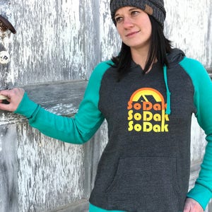 May include: A charcoal grey hooded sweatshirt with teal green sleeves and trim. The front of the sweatshirt features a graphic of a sunset with the text "SoDak" repeated three times in a retro font.