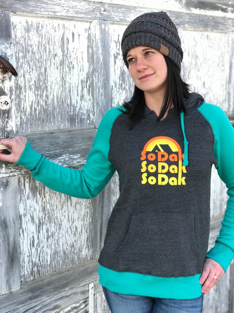 May include: A charcoal grey hooded sweatshirt with teal green sleeves and trim. The front of the sweatshirt features a graphic of a sunset with the text "SoDak" repeated three times in a retro font.