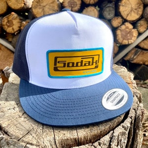 May include: White and navy blue trucker hat with a yellow and turquoise patch that says "Sodak" in brown lettering. The hat has a snap closure.