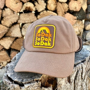 Puede incluir: Una gorra de béisbol marrón con un parche que presenta una cordillera amarilla y naranja y el texto "SoDak" repetido tres veces.