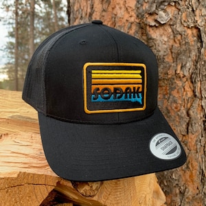 May include: Black trucker hat with a yellow, orange, and blue striped patch that says "SODAK" in a retro font.