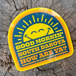 May include: A yellow and blue sticker with a smiling sun and the text "Good Morning' South Dakota How Are Ya?"  on a wood background. The sticker has a retro design.