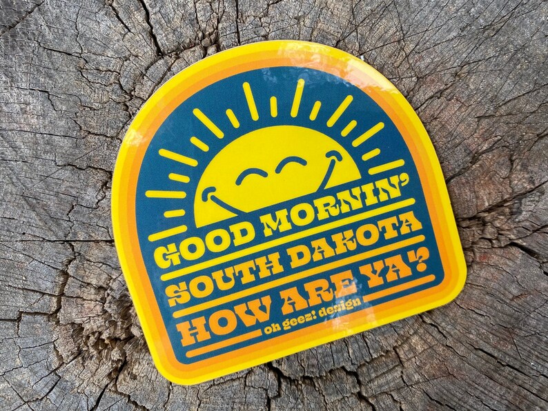 May include: A yellow and blue sticker with a smiling sun and the text "Good Morning' South Dakota How Are Ya?"  on a wood background. The sticker has a retro design.