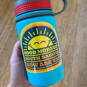 May include: A blue water bottle with a gray lid and a yellow and orange sticker that says "Good Morning South Dakota How Are Ya!" with a smiley face sun graphic. The sticker also says "oh geez! design".