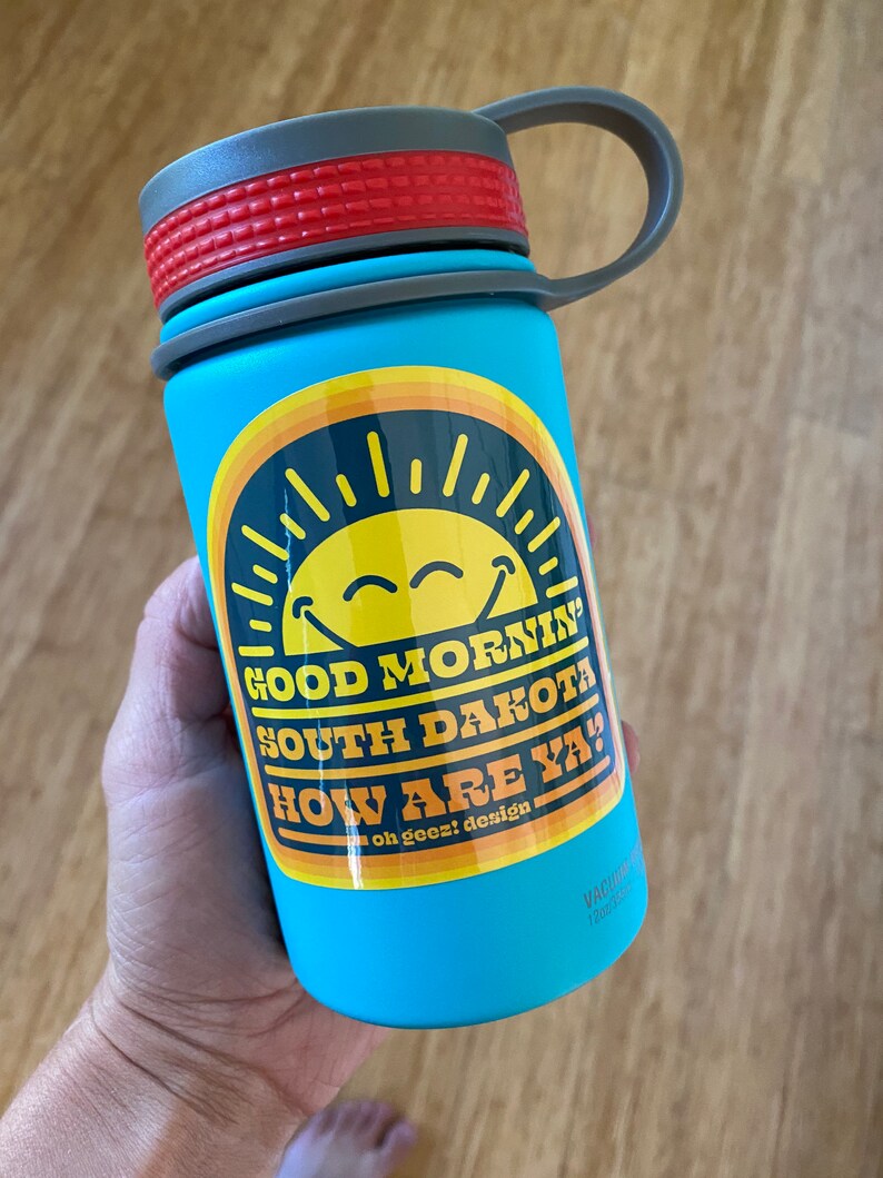 May include: A blue water bottle with a gray lid and a yellow and orange sticker that says "Good Morning South Dakota How Are Ya!" with a smiley face sun graphic. The sticker also says "oh geez! design".