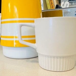May include: A white ceramic mug with a ribbed base and a yellow and white striped jug in the background.