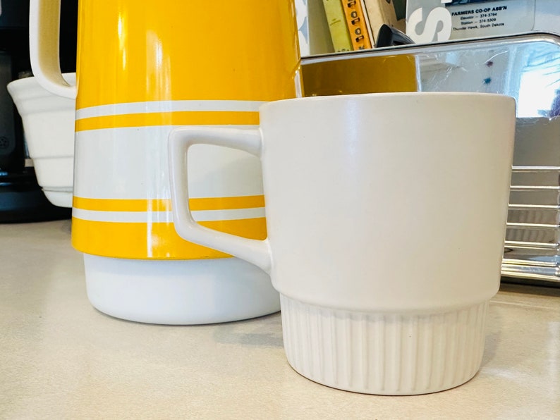 May include: A white ceramic mug with a ribbed base and a yellow and white striped jug in the background.