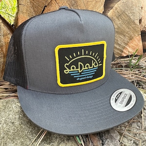 May include: A gray and black trucker hat with a yellow and black patch that says "So Dak" with a sunset and waves design. The patch also says "oh great design".