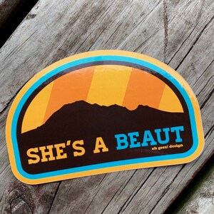 May include: A yellow and blue sticker with a silhouette of a mountain range and the text "SHE'S A BEAUT oh geez! design".