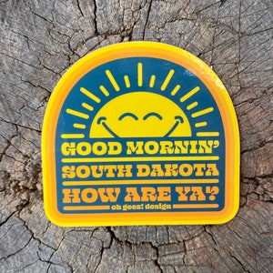 May include: Yellow and blue sticker with a smiling sun and the text "Good Mornin' South Dakota How Are Ya?" and "oh geez! design"