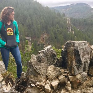 May include: A woman wearing a teal and black hooded sweatshirt with the text "SoDak SoDak SoDak" in a retro font. She is standing on a rocky cliff overlooking a forest.