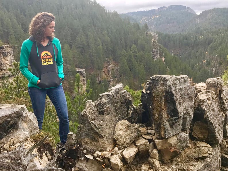 May include: A woman wearing a teal and black hooded sweatshirt with the text "SoDak SoDak SoDak" in a retro font. She is standing on a rocky cliff overlooking a forest.