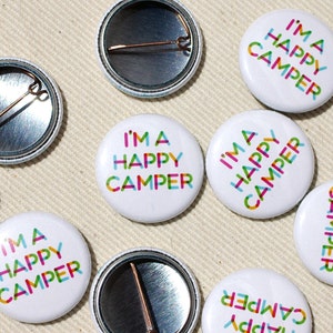 May include: A set of eight round white button badges with a colourful graphic design that reads "I'm a Happy Camper".