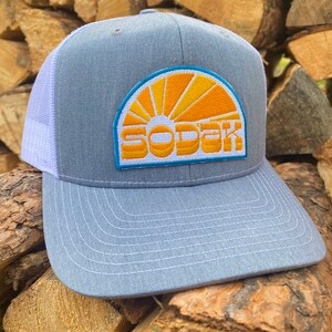 May include: A gray and white trucker hat with a white mesh back. The front of the hat features a yellow and orange embroidered patch with a sun and the word "SODAK".