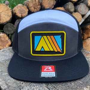 May include: A black, gray, and light blue trucker hat with a yellow and orange patch featuring a stylized tent and the text "WEAR THE GEAR". The hat has a black mesh back and a black flat bill with a white "Richardson" label that reads "FLAT BILL SNAPBACK".