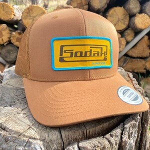 May include: A brown trucker hat with a yellow and blue patch that says "Sodak" on it. The hat has a mesh back and a curved brim.