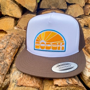 May include: A brown and white trucker hat with a yellow and orange sunrise graphic and the word "SODAK" in blue and white lettering. The hat has a brown mesh back and a brown brim. The hat has a white label on the back that says "The Authentic Yupoong Snapback Since 1974."