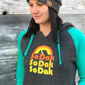 May include: A dark gray hooded sweatshirt with teal green sleeves. The front of the sweatshirt has a graphic design of a rainbow with a tent inside and the text "So Dak" repeated three times.
