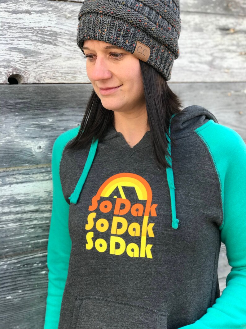 May include: A dark gray hooded sweatshirt with teal green sleeves. The front of the sweatshirt has a graphic design of a rainbow with a tent inside and the text "So Dak" repeated three times.