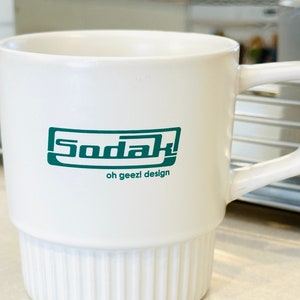 May include: White ceramic mug with a green logo that says "Sodak! oh geez! design".