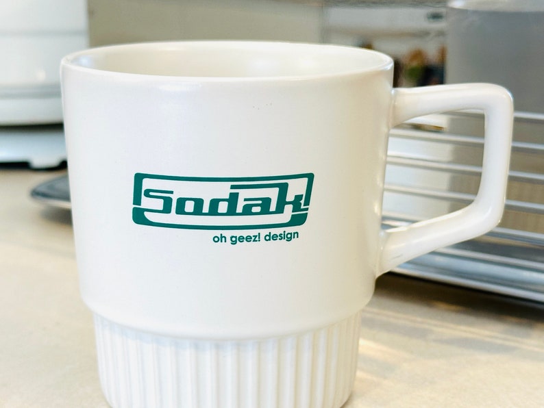 May include: White ceramic mug with a green logo that says "Sodak! oh geez! design".