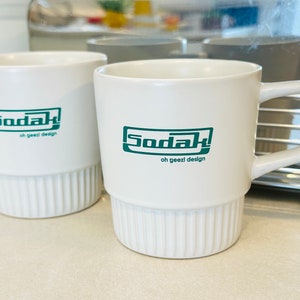 May include: Two white ceramic mugs with a green logo that says "Sodak oh geezi design".