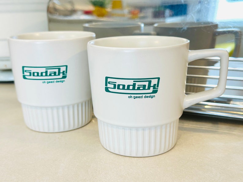 May include: Two white ceramic mugs with a green logo that says "Sodak oh geezi design".