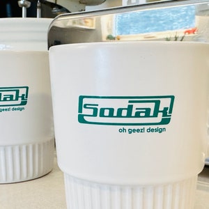 May include: Two white ceramic mugs with a green logo that says "Sodak oh geezl design".