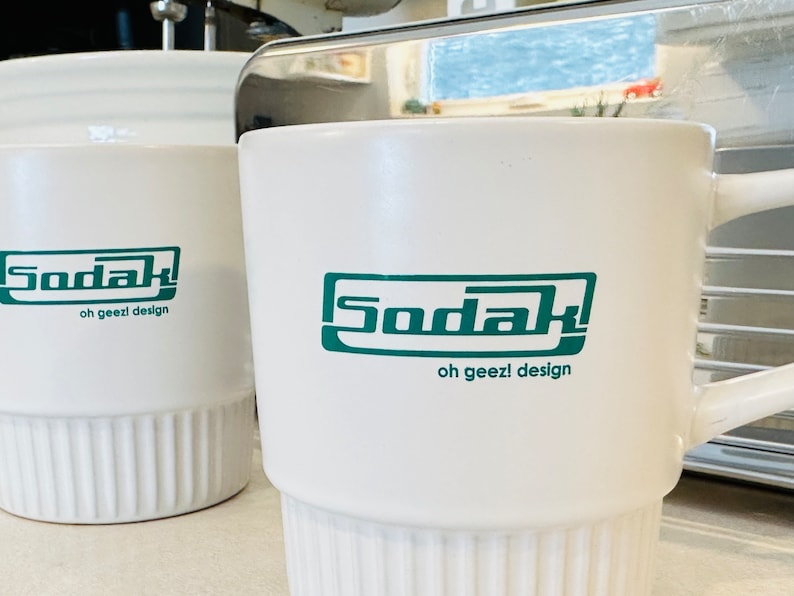 May include: Two white ceramic mugs with a green logo that says "Sodak oh geezl design".