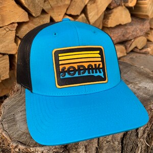 May include: A blue and black trucker hat with a yellow, orange, and black patch that says "SODAK" on it. The patch has a retro sunset design.