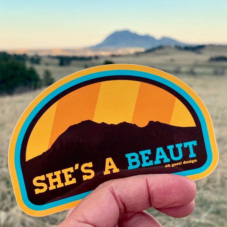 May include: A yellow and blue sticker with a silhouette of a mountain range and the text "SHE'S A BEAUT oh geez! design".
