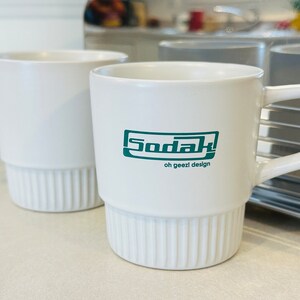 May include: Two white ceramic mugs, one with the text "Sodak! oh geezi design" in green lettering.