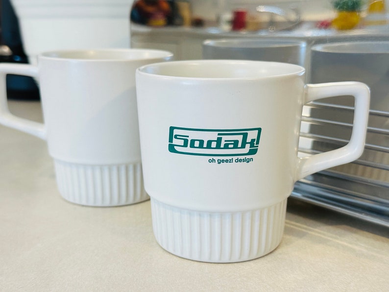 May include: Two white ceramic mugs, one with the text "Sodak! oh geezi design" in green lettering.