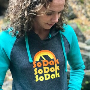 May include: A dark gray hooded sweatshirt with teal sleeves. The front of the sweatshirt has a retro-style graphic of a mountain range with a rainbow and the text "So Dak" repeated three times.