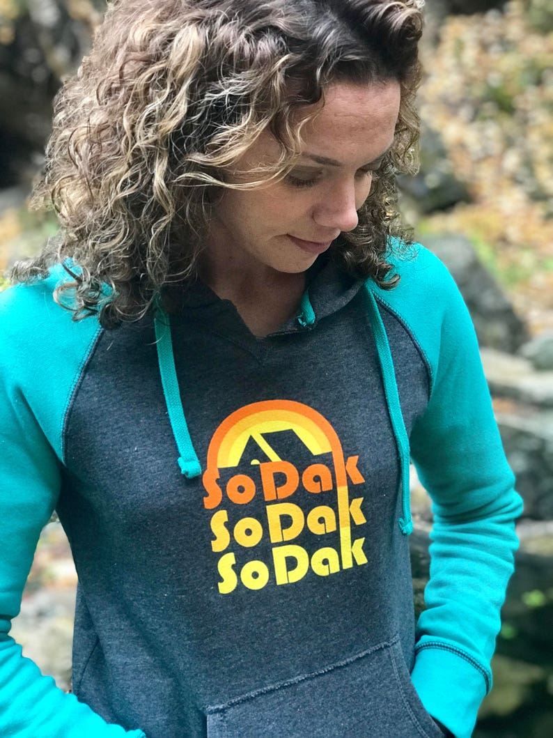 May include: A dark gray hooded sweatshirt with teal sleeves. The front of the sweatshirt has a retro-style graphic of a mountain range with a rainbow and the text "So Dak" repeated three times.