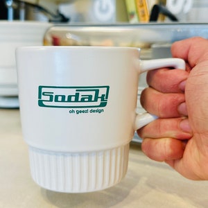 May include: White ceramic mug with a green logo that says "Sodak! oh geezl design"