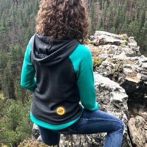 May include: A woman wearing a teal and gray hooded sweatshirt with a yellow sun graphic on the back. She is standing on a rocky cliff overlooking a forest.
