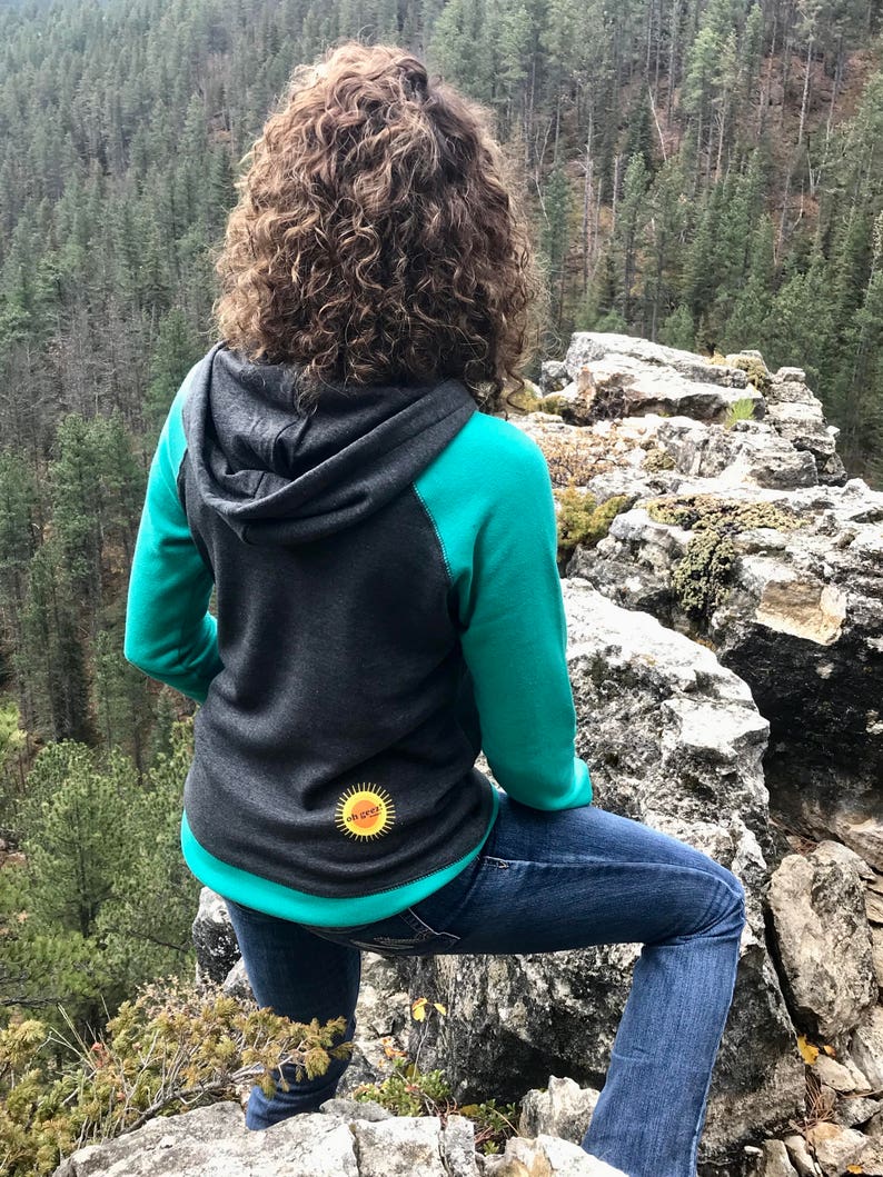 May include: A woman wearing a teal and gray hooded sweatshirt with a yellow sun graphic on the back. She is standing on a rocky cliff overlooking a forest.