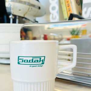 May include: White ceramic mug with a green logo that says "Sodak" and "oh geezl design".