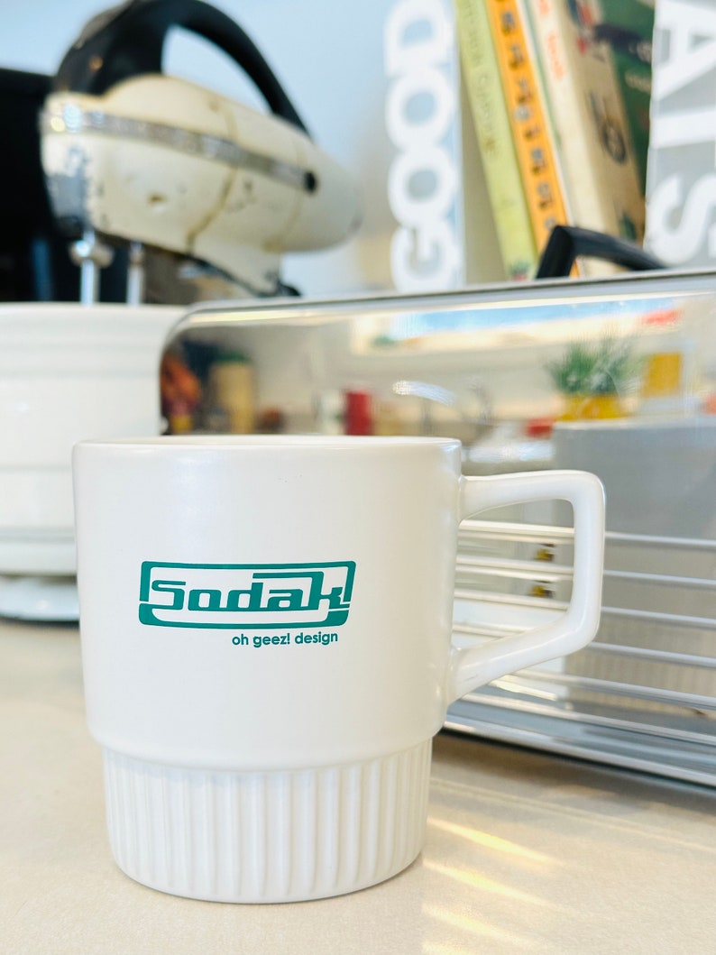 May include: White ceramic mug with a green logo that says "Sodak" and "oh geezl design".