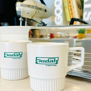 May include: Two white ceramic mugs with green text that reads "Soda! oh geez! design". The mugs have a ribbed design and are sitting on a counter.