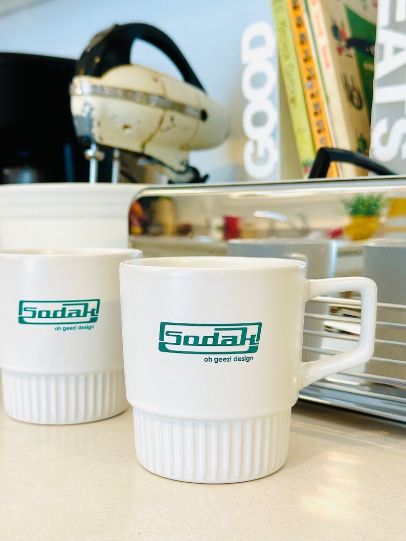 May include: Two white ceramic mugs with green text that reads "Soda! oh geez! design". The mugs have a ribbed design and are sitting on a counter.