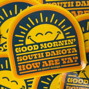 May include: A yellow and orange sticker with a smiling sun and the text "Good Mornin' South Dakota How Are Ya?" with the tagline "oh geez! design".