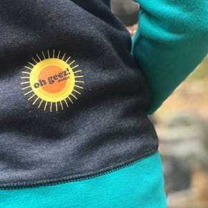 May include: A dark gray sweatshirt with a teal green trim. The sweatshirt has a yellow sun with the text "oh geez!" in orange on the front.