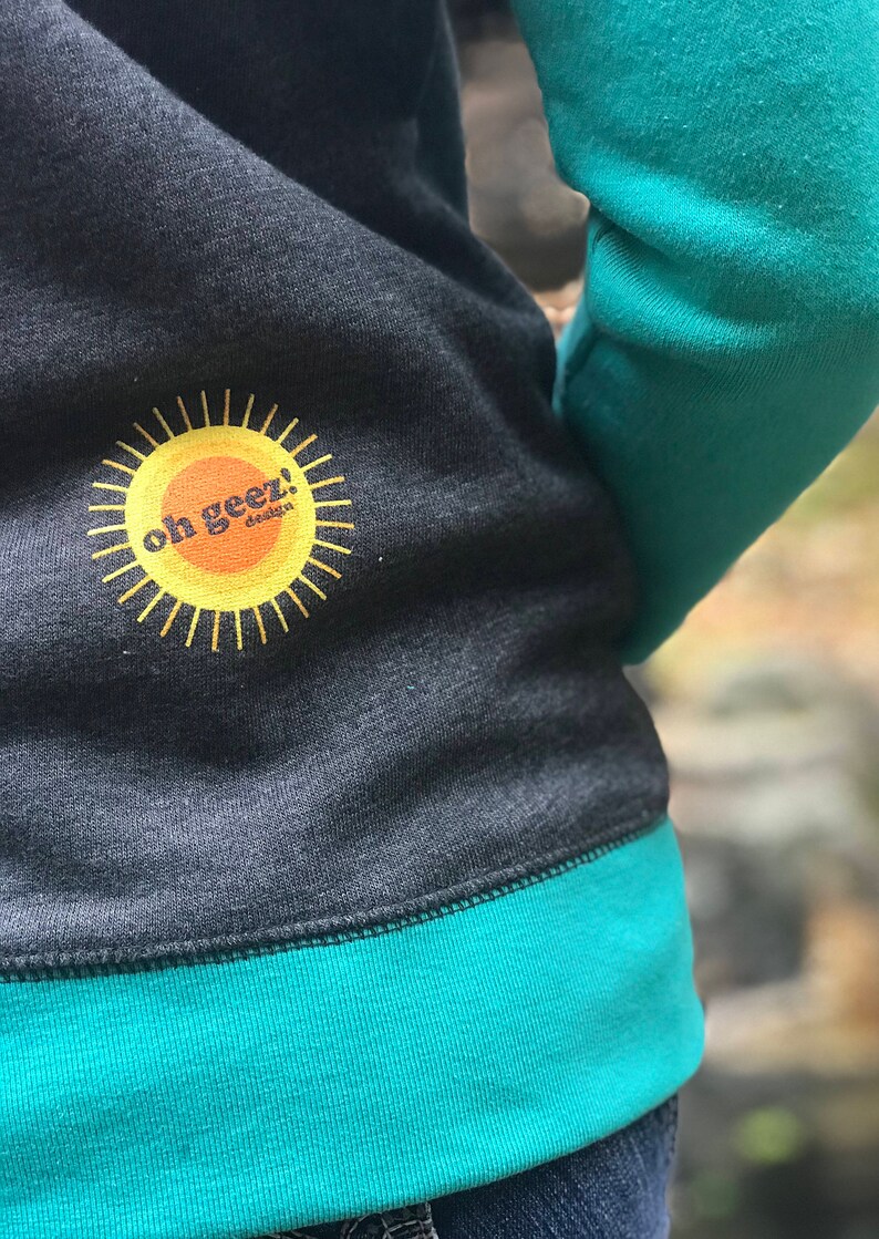 May include: A dark gray sweatshirt with a teal green trim. The sweatshirt has a yellow sun with the text "oh geez!" in orange on the front.