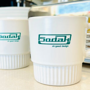 May include: Two white ceramic mugs with a green logo that says "Sodak oh geez! design". One mug has a handle and the other does not.